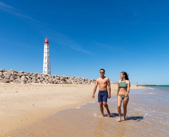 Fotografia do Farol visto da praia, vemos também um casal a passear a beira mar
